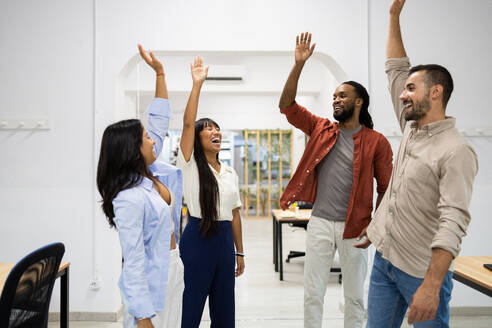 Cheerful successful multiracial male and female coworkers in casuals raising hands together while standing in creative office - ADSF51275