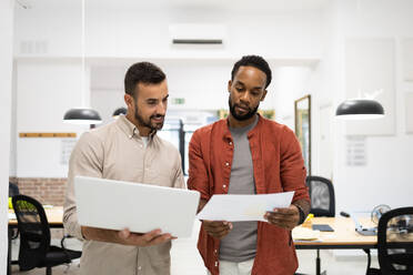 Multiracial male colleagues in discussing reports over document and laptop while standing in creative office - ADSF51276