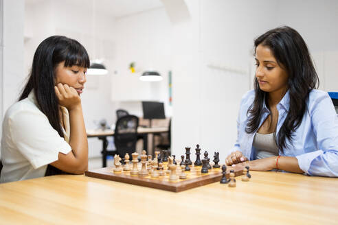 Serious multiracial young female colleagues dressed in casuals playing chess while sitting at wooden desk in creative office - ADSF51278