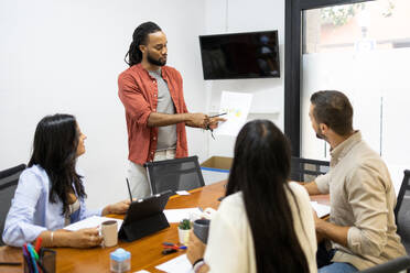 Confident businessman explaining multiracial male and female coworkers over document during meeting in creative office - ADSF51279