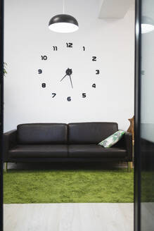 Creative black clock on white wall over empty sofa in lobby of coworking office seen through doorway - ADSF51285