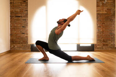 A man performs a yoga stretch on a mat in a room with hardwood floors and exposed brick walls, illuminated by natural light. - ADSF51292