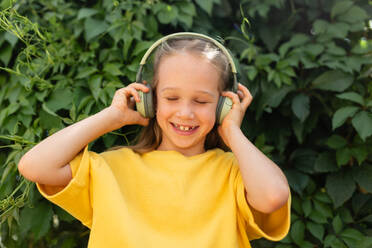 A cheerful young girl in a yellow dress listens to music through green headphones against a leafy backdrop. - ADSF51308