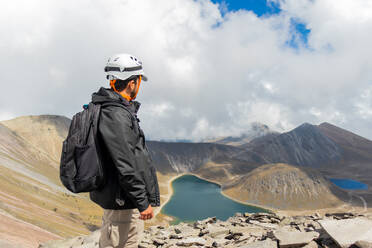 A hiker in safety gear stands atop a mountain, overlooking the stunning lakes nestled within Nevado de Toluca's craters under a cloudy sky. - ADSF51310