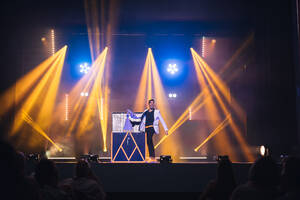 Magician performing magic trick on dark stage with glowing orange spotlights while taking stuff out of big box during performance - ADSF51316