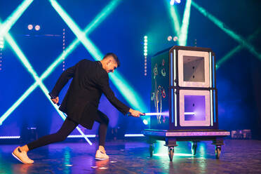 Side view of magician in black suit performing magic trick with boxes on dark stage illuminated by blue and green spotlights in theater - ADSF51317