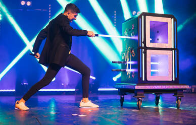 Side view of magician in black suit performing magic trick with boxes on dark stage illuminated by blue and green spotlights in theater - ADSF51318