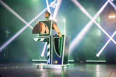 Male magician showing magic trick with half body in box on stage illuminated by spotlights during performance - ADSF51319