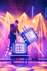 Professional male magician on top of a table showing magic trick with glass boxes and swords on stage with smoke and glowing spotlights during performance - ADSF51320