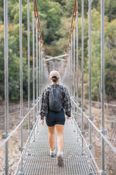 Back view of anonymous female hiker in shorts and backpack walking on narrow footbridge while trekking in forest during weekend holiday - ADSF51321