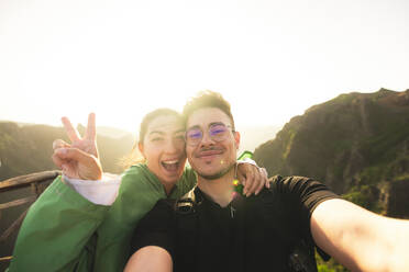 Portrait of smiling Caucasian young man taking selfie with cheerful woman showing victory sign while standing on top of mountain against clear sky - ADSF51327