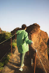 Back view of young unrecognizable woman dressed in green raincoat walking on trail by railing at mountain peak against clear sky during sunrise - ADSF51332