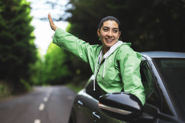 Happy young woman with arms outstretched looking away and leaning out of car window while enjoying road trip in forest - ADSF51333