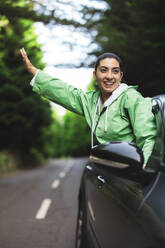 Happy young woman with arms outstretched looking away and leaning out of car window while enjoying road trip in forest - ADSF51334