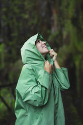 Smiling young woman wearing green raincoat with hoodie looking up while hiking by cliff covered in moss during vacation - ADSF51346