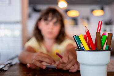 Various color pencils and pens in plastic cup placed on wooden table against caucasian elementary girl spending leisure time at restaurant - ADSF51357