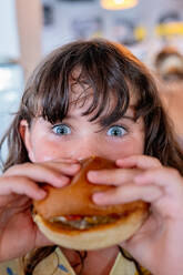 Closeup hands of hungry anonymous girl holding and eating delicious hamburger and looking at camera while spending leisure time in cafe - ADSF51359