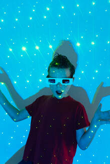 Portrait of Cute boy in casual clothes with beads on face and 3D glasses standing with opened mouth against wall with illuminated blue neon lights while looking at camera - ADSF51361