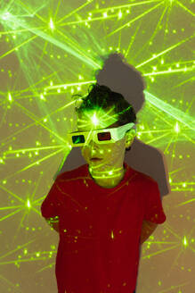 High angle of Cute boy in casual clothes with beads on face and 3D glasses standing against wall with illuminated yellow neon lights while looking away - ADSF51362