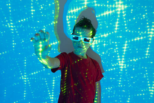 Portrait of Cute boy in casual clothes with beads on face and 3D glasses standing with hand out against wall with illuminated blue neon lights while looking at camera - ADSF51363