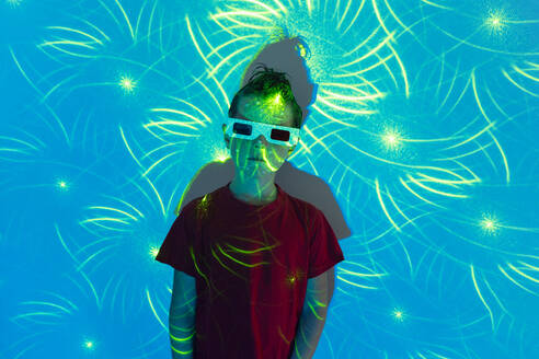 Portrait of Cute boy in casual clothes with beads on face and 3D glasses standing against wall with illuminated blue neon lights while looking at camera - ADSF51364