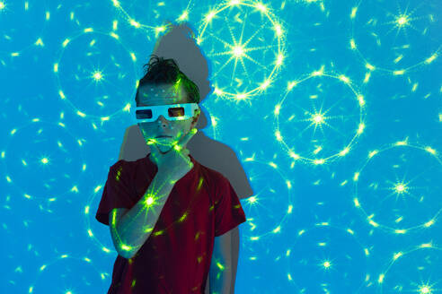 Portrait of Cute boy in casual clothes with beads on face and 3D glasses standing against wall with illuminated blue neon lights while looking at camera and touching chin - ADSF51365