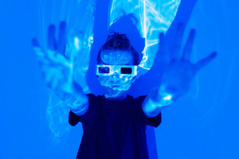 Portrait of Confident boy in casual clothes with beads on face and 3D glasses standing with hands out in illuminated blue neon studio against wall while looking at camera - ADSF51366