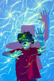 Amazed boy wearing virtual reality goggles with raised arms and open mouth experiencing cyberspace while standing in studio with neon lights - ADSF51367