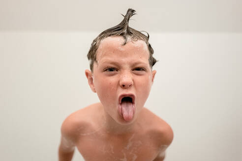 Little boy with foam on head and skin looking at camera while bathing in bathroom and showing tongue out against white background - ADSF51371