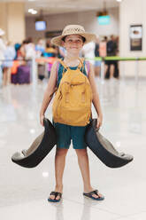 A smiling young boy in shorts and a straw hat grips large suitcase handles in an airport - ADSF51372