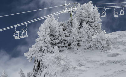 Low angle view of ropeway with empty chairlifts above snow covered trees and mountain against cloudy sky in winter nature - ADSF51397