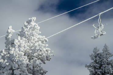 Low angle view of ropeway with empty chairlifts above snow covered trees and mountain against cloudy sky in winter nature - ADSF51398