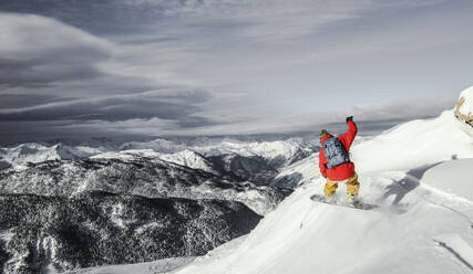 Back view of anonymous carefree person with backpack snowboarding on snowcapped mountain top against majestic view of mountains and sky during vacation - ADSF51403