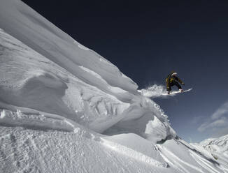 Low angle view of adventures unrecognizable snowboarder jumping from snow covered rocky mountain against blue sky during winter vacation on sunny day - ADSF51410