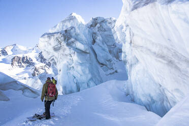 Back view of anonymous hiker with backpack snowboarding on snow-covered mountain near icy peaks in Zermatt, Switzerland with friend under blue sky - ADSF51429