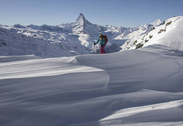 Side view of Unrecognizable female backpacker in warm clothes and ski walking on snowy landscape against majestic rocky mountain in Zermatt, Switzerland under blue sky - ADSF51438