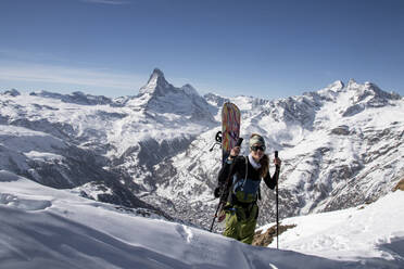 Smiling Young female traveler in warm clothes with snowboard holding ski poles while looking at camera and standing against snowy rocky mountains in Zermatt, Switzerland under blue sky - ADSF51442