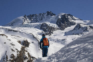 Back view of anonymous male backpacker standing with ski poles on snowy mountain under blue sky during vacation in Zermatt, Switzerland - ADSF51452