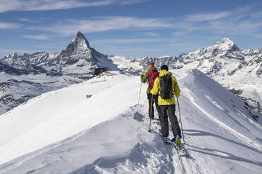 Back view of anonymous men in warm clothes skiing on snow-covered landscape against majestic rocky mountains under cloudy sky in Zermatt, Switzerland - ADSF51453