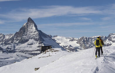 Back view of anonymous men in warm clothes skiing on snow-covered landscape against majestic rocky mountains under cloudy sky in Zermatt, Switzerland - ADSF51454