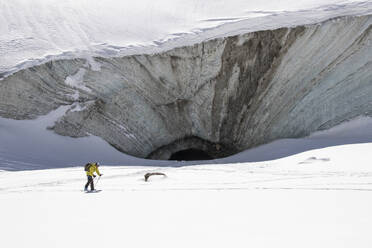 Side view of anonymous tourist with backpack skiing while approaching the gaping maw of a glacier cavern on sunny day in Zermatt, Switzerland - ADSF51457