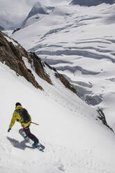 High angle of anonymous Male Tourist with backpack snowboarding on majestic snow covered mountain slope against mountains under cloudy sky in Zermatt, Switzerland - ADSF51458