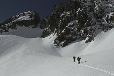 Back view of unrecognizable friends in warm clothes with backpacks skiing on snow covered landscape against beautiful rocky mountains on sunny day at Swiss Alps - ADSF51474