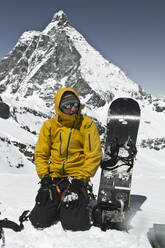 Male snowboarder wearing warm clothes and sunglasses kneeling with snowboard on snow covered landscape against mountain peak during winter holidays at Swiss Alps - ADSF51479