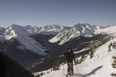 Anonymous active skier with poles and backpack skiing on snow covered mountain slope under clear sky during sunny day in Canada - ADSF51487