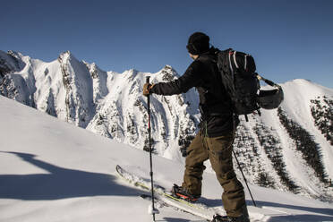 Side view of anonymous active skier with poles and backpack skiing on snow covered mountain slope under clear sky during sunny day in Canada - ADSF51491