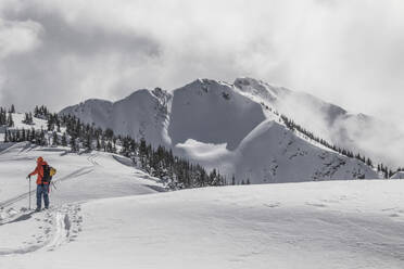 Back view of anonymous active skier with poles and backpack skiing on snow covered mountain slope under cloudy sky during sunny day in Canada - ADSF51494