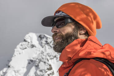 Side view of male traveler in orange warm clothes and goggles standing against snowcapped mountain peak and clear sky during winter vacation on sunny day - ADSF51496