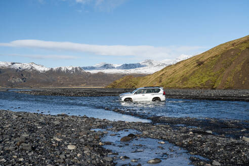 An adventurous off-road SUV vehicle fords through a pristine river against a scenic backdrop of the rugged Icelandic highlands under blue sky with clouds - ADSF51521