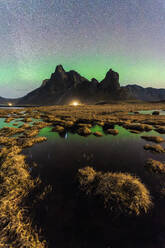A stunning view of the aurora borealis dancing over a craggy mountain peak, mirrored in a tranquil pond below under a starry sky Eystrahorn, Iceland - ADSF51569